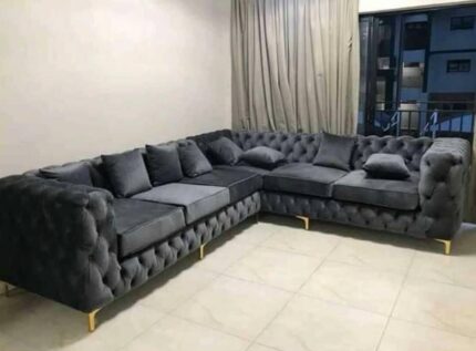 “L-shaped button couch in grey fabric with deep tufting, plush cushions, and a sturdy wooden frame, placed in a modern living room setting.”
