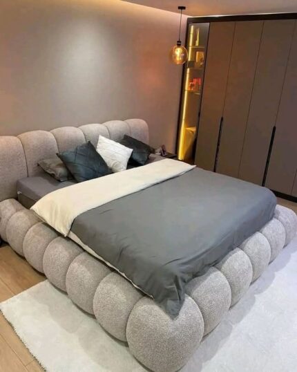 A luxurious modern bed from King’s Touch Furniture featuring a unique bubble-style upholstered base and a curved padded headboard. The bed is dressed in gray and beige bedding, placed in a stylish bedroom with warm lighting and a sleek wardrobe in the background.