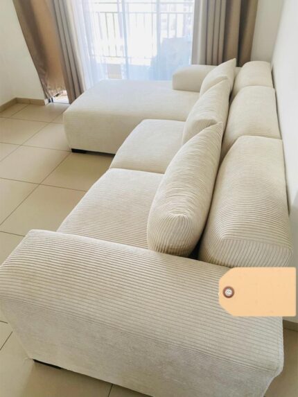 A modern L-shaped sectional couch upholstered in light beige corduroy fabric,placed in a bright living room with tiled floors and sheer curtains letting in natural light.