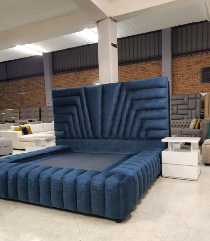 A luxurious blue upholstered bed by King’s Touch Furniture, featuring a tall geometric-patterned headboard and matching padded frame, displayed in a modern furniture showroom with white side tables and other furniture pieces in the backg