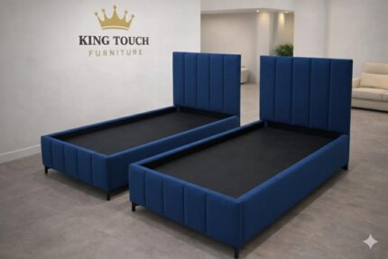 Two matching royal blue velvet Vertical Channel Single Beds by King Touch Furniture, featuring high-profile headboards with vertical panel tufting and coordinating upholstered bases with sleek black legs, displayed in a minimalist showroom.